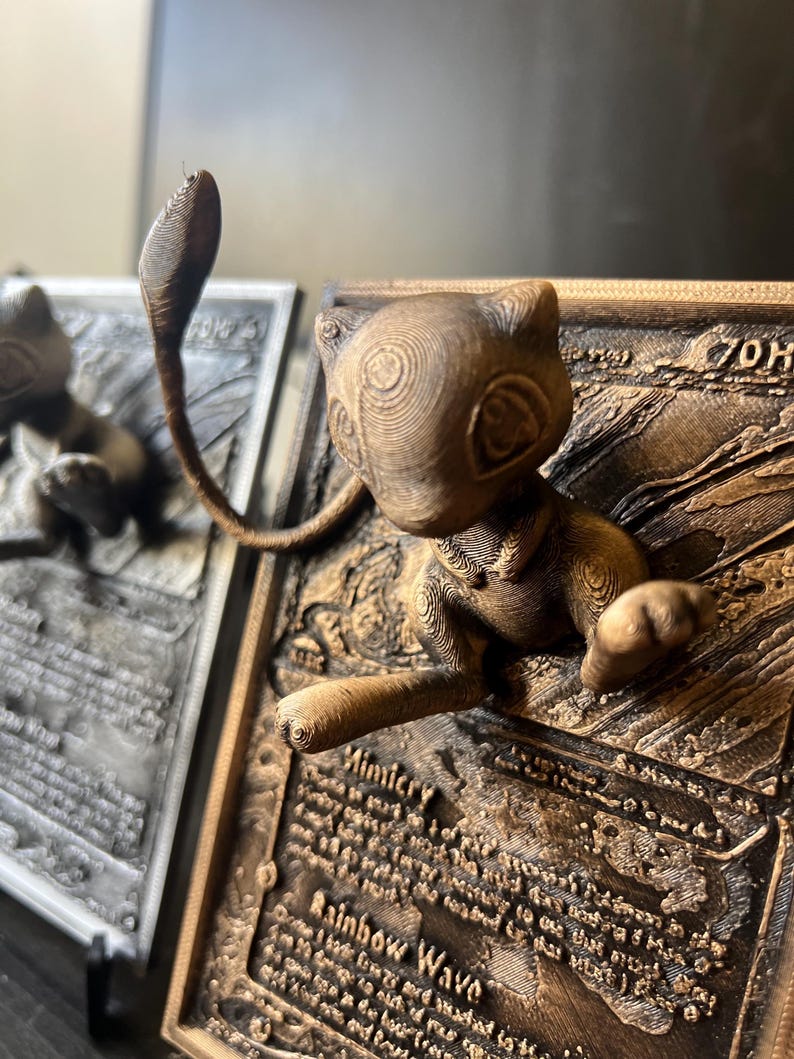 May include: A 3D printed bronze-colored Pokemon card featuring a detailed sculpture of the Pokemon, Mewtwo, with its signature psychic powers. The card is titled "Rainbow Wave" and includes the text "Mimicry".