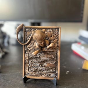 May include: A bronze 3D printed Pokemon card featuring Mew, a Psychic-type Pokemon. The card has a brown background with gold accents and features the text "Mew" and "70 HP". The card also has the text "Mimicry" and "Rainbow Wave" on it.