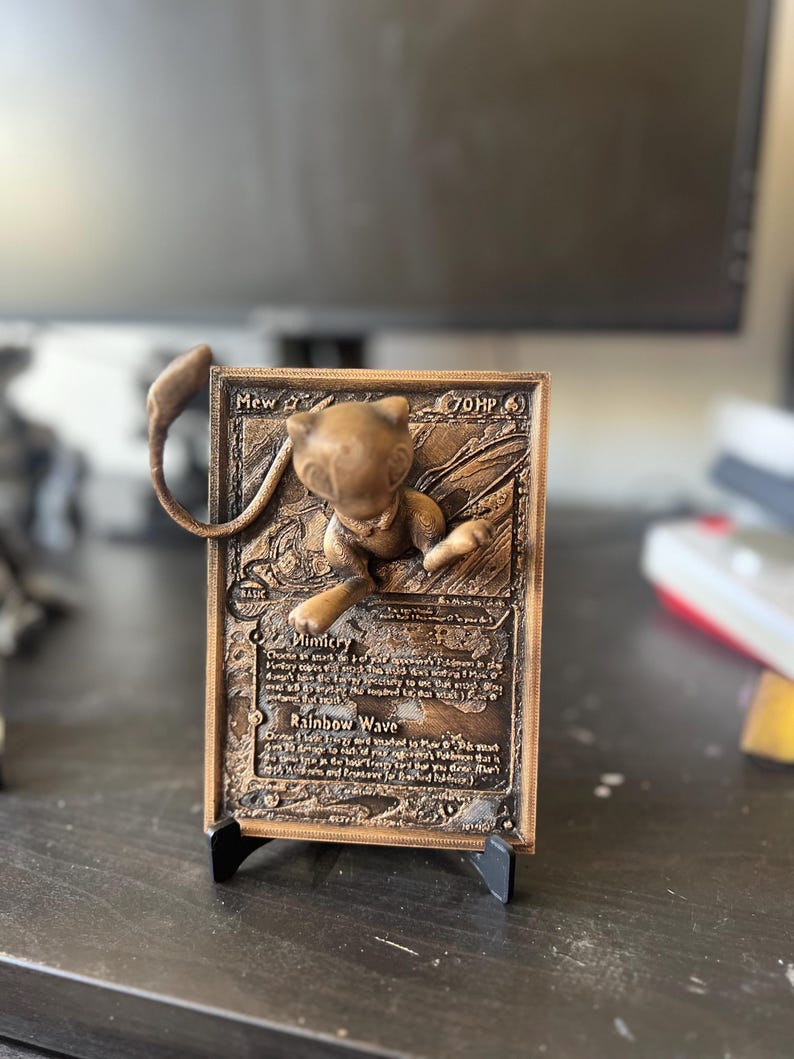 May include: A bronze 3D printed Pokemon card featuring Mew, a Psychic-type Pokemon. The card has a brown background with gold accents and features the text "Mew" and "70 HP". The card also has the text "Mimicry" and "Rainbow Wave" on it.