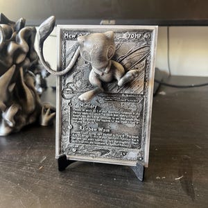 May include: A 3D printed Pokemon card featuring Mew, a pink and white mythical Pokemon. The card is silver and has a black stand. The card text reads "Mew - 70 HP" and includes the moves "Mimicry" and "Rainbow Wave".