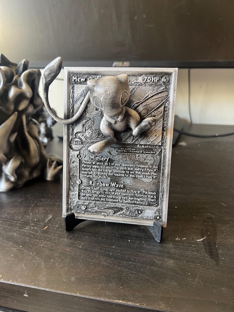 May include: A 3D printed Pokemon card featuring Mew, a pink and white mythical Pokemon. The card is silver and has a black stand. The card text reads "Mew - 70 HP" and includes the moves "Mimicry" and "Rainbow Wave".