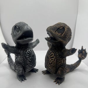 May include: Two small, detailed figurines of a mythical creature with a lizard-like body and a dragon-like head. The figurine on the left is painted in a silver color, while the figurine on the right is painted in a bronze color.