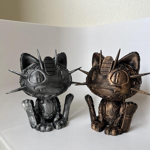 May include: Two cat figurines with a steampunk aesthetic. One is silver, the other bronze. Both have spiky protrusions and intricate detailing. They are sitting with their legs outstretched.