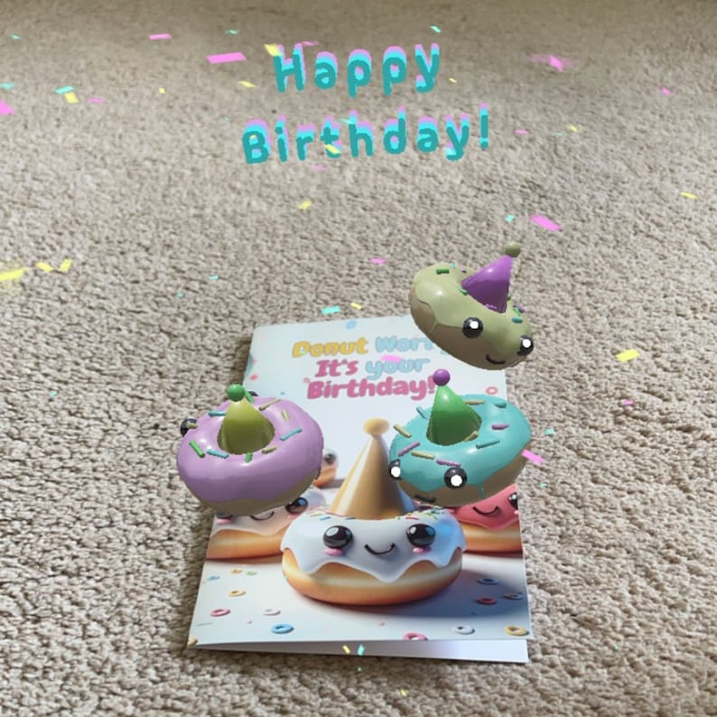 May include: A birthday card with the text "Donut Worry It's Your Birthday!" and three colorful donuts with happy faces and party hats. The donuts are floating above the card, which is on a beige surface with confetti.