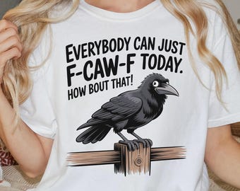 Pode incluir: Camiseta branca com um desenho de um corvo preto empoleirado numa viga de madeira. O texto diz: "EVERYBODY CAN JUST F-CAW-F TODAY. HOW BOUT THAT!" Parte de uma coleção com tema de outono.