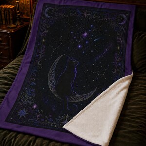 Puede incluir: Una manta rectangular con borde morado oscuro y reverso de forro polar blanco. El frente muestra un gato negro sentado sobre una luna creciente en un cielo estrellado, rodeado por un marco ornamentado con motivos florales y celestiales.