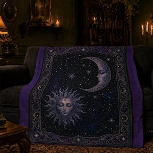 May include: A rectangular throw blanket featuring a dark navy and purple celestial design. The center displays a stylized sun and crescent moon with human faces, surrounded by stars and constellations. A decorative border frames the imagery.