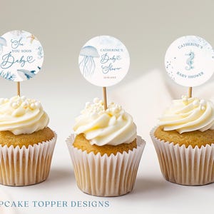 May include: Three cupcakes with white frosting and decorative toppers. The toppers feature aquatic designs and text that reads "Sea You Soon Baby", "Catherine's Baby Shower", and "Baby Shower". The cupcakes are in white paper liners.