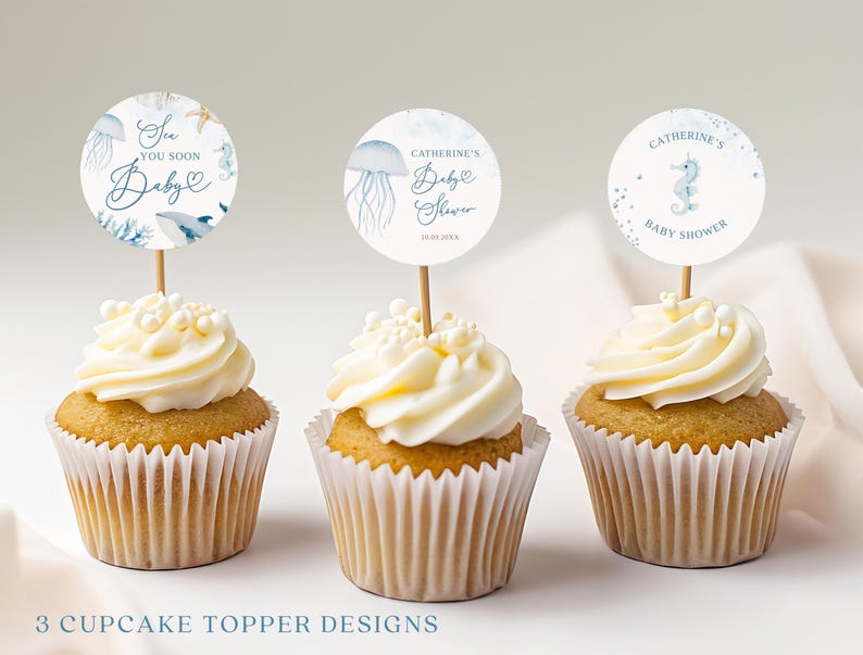 May include: Three cupcakes with white frosting and decorative toppers. The toppers feature aquatic designs and text that reads "Sea You Soon Baby", "Catherine's Baby Shower", and "Baby Shower". The cupcakes are in white paper liners.