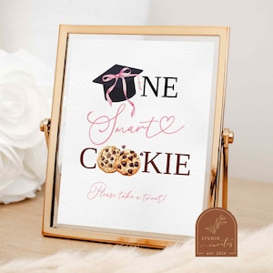 Puede incluir: Un cartel con marco dorado con el texto "One Smart Cookie" en letras negras y rosas. El cartel también presenta un gorro de graduación con una cinta rosa y dos galletas de chocolate.