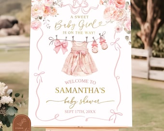 May include: A pink and gold baby shower welcome sign with floral accents and a pink dress hanging on a clothesline. The text reads "A Sweet Baby Girl is on the way! Welcome to Samantha's Baby Shower Sept 17th, 20XX".