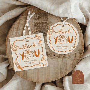 Może przedstawiać: Dwie kremowe papierowe zawieszki prezentowe z pomarańczowymi rysunkami liniowymi dyń, jabłek i napojów. Tekst brzmi Thank YOU, Anna's Birthday oraz Oct 12, 20xx. Jedna zawieszka jest kwadratowa, druga okrągła z falistym brzegiem.