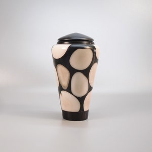 Peut inclure: Un urne décorative avec un couvercle et une base noirs, présentant un motif d'ovales crème sur un fond noir. L'urne a une forme conique et est probablement en bois ou dans un matériau similaire.