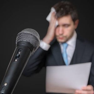 Może przedstawiać: Czarny mikrofon z siatką w kolorze szarym znajduje się na pierwszym planie. Osoba w garniturze jest rozmyta w tle, trzymając kartkę papieru i wycierając czoło białym ręcznikiem.