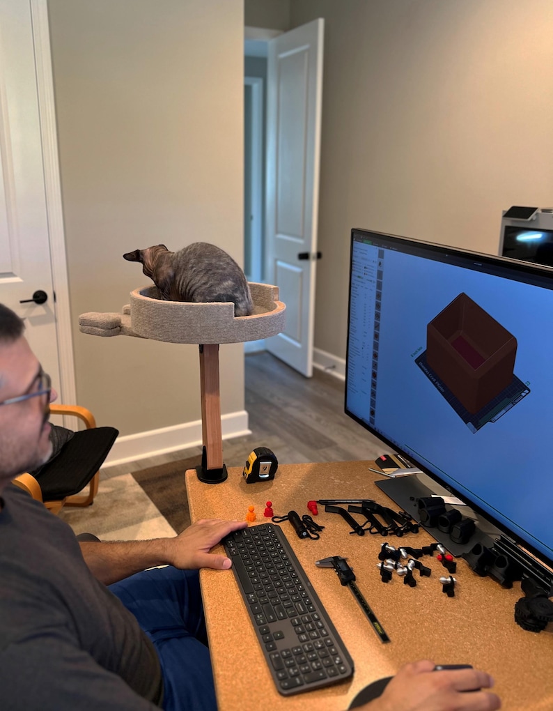 Op de afbeelding: Een grijze kat zit op een beige kattenboom met een houten paal. Een persoon zit aan een bureau met een computermonitor waarop een 3D-ontwerp van een doos te zien is. Het bureau heeft een kurk oppervlak en een toetsenbord, gereedschap en andere items.