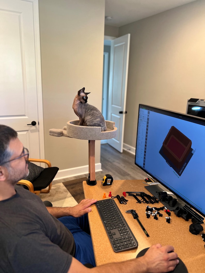 Op de afbeelding: Een persoon zit aan een bureau met een kurk oppervlak. Een zwart toetsenbord, een meetlint en verschillende gereedschappen liggen op het bureau. Een computermonitor toont een technische tekening. Een kat zit op een kattenboom op de achtergrond.