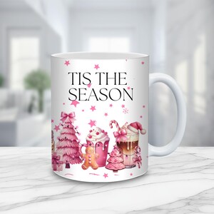 May include: A white ceramic mug featuring the black text 'TIS THE SEASON' surrounded by pink stars and holiday illustrations including a pink Christmas tree, a gingerbread man, a pink mug with whipped cream, and a coffee drink with a candy cane.