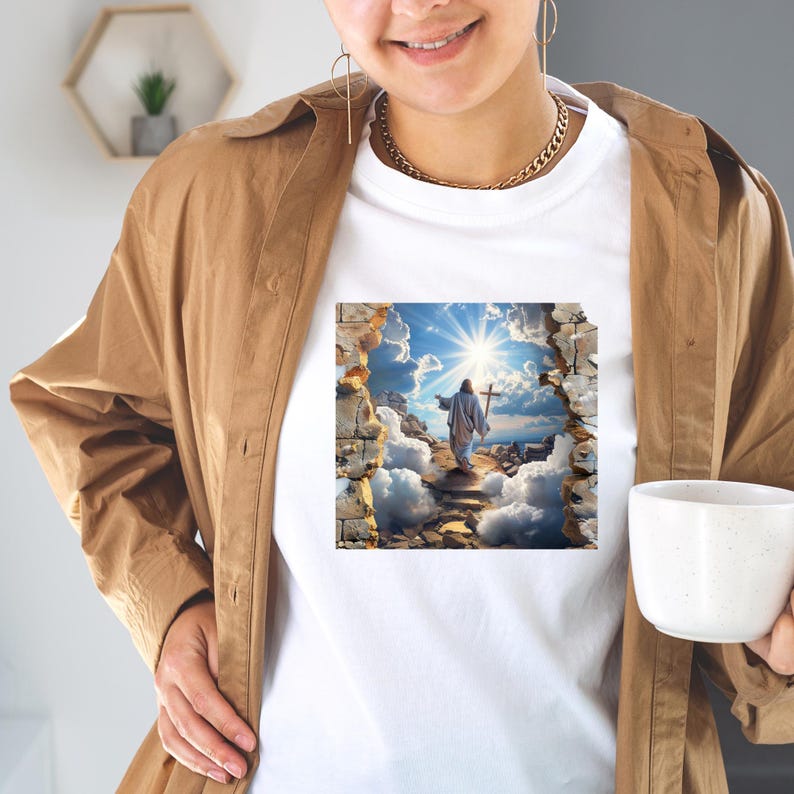 May include: A white t-shirt featuring a graphic print of a figure holding a cross, walking toward a bright sunlit sky through a stone wall opening. The shirt is worn under a tan button-down shirt, with a gold chain necklace and a white mug.