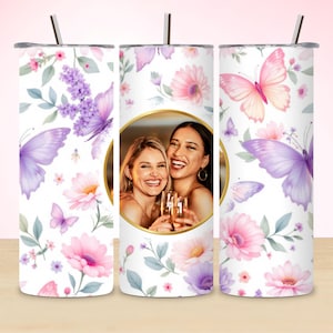 May include: A white tumbler with a floral and butterfly design. The design features pink and purple flowers, lavender sprigs, and butterflies. A gold-rimmed oval in the center holds a photo of two smiling women holding champagne flutes. A black straw is inserted in the lid.