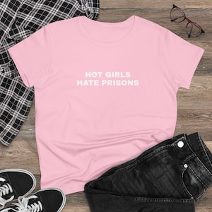 Puede incluir: Una camiseta rosa claro con el texto blanco "HOT GIRLS HATE PRISONS". La camiseta se muestra con vaqueros negros, zapatillas deportivas blancas y negras y gafas.