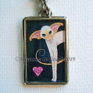 Può includere: Un ciondolo d'argento con un gatto siamese bianco e arancione con occhi azzurri e un cuore rosa su sfondo nero. Il ciondolo è incorniciato in argento e ha il testo "Suzanne Le Good 2019" in basso.