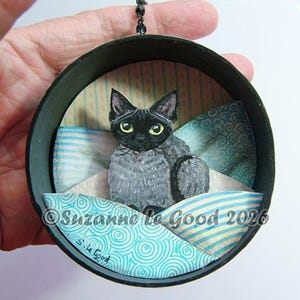 Könnte beinhalten: Rundes Kunstwerk mit schwarzem Rahmen, das eine graue Katze mit gelben Augen auf blau-weiß gemusterten Kissen zeigt. Der Text "Suzanne le Good 2026" und "S. le Good" sind zu sehen.