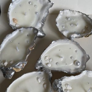 May include: A collection of oyster shell candles. Each candle has creamy white wax, small seashells, and faux pearls. The shells have a silver metallic trim. Each candle has a single wick.