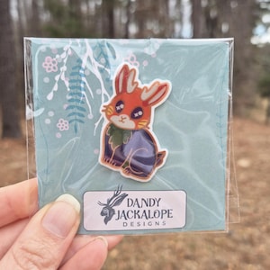 May include: A colorful sticker featuring a jackalope design with orange, purple, and green accents. The sticker is on a light blue background with floral and leaf patterns. The sticker is in a clear plastic sleeve, with the text "DANDY JACKALOPE DESIGNS" on a white label.