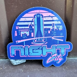 May include: A blue sticker with a retro design featuring a cityscape silhouette in pink and blue. The word "NIGHT" is in large, block letters, and "City" is in a cursive font. The sticker has a distressed, vintage look.