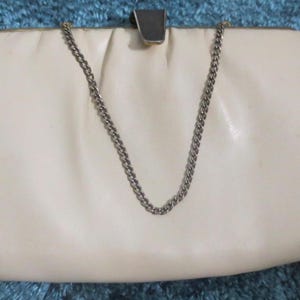 May include: Cream-colored clutch purse with a silver chain strap. The purse has a rectangular shape with a curved top and a metal clasp. The chain is a silver color and hangs down from the top of the purse.