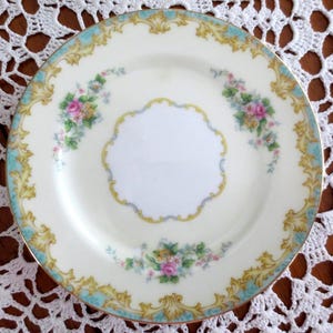 May include: A cream-colored porcelain plate with a floral design. The plate features a light blue border with gold filigree and pink and yellow floral clusters. The center has a white space with a decorative yellow and blue border.