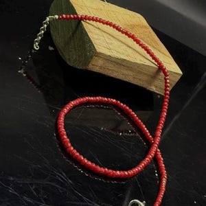 Op de afbeelding: Een delicate rode kralenketting met een zilveren sluiting. De ketting bestaat uit kleine, gefacetteerde rode kralen die aan elkaar zijn geregen. De ketting wordt gepresenteerd op een donkere ondergrond met een houten blok op de achtergrond.