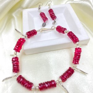 May include: A jewelry set featuring a bracelet and earrings. The bracelet and earrings are made of faceted, deep red beads and silver-toned metal accents. The earrings have a leaf charm. The bracelet has feather charms.