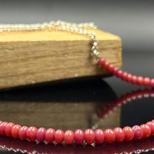 May include: A necklace featuring small, round, red beads strung on a silver chain. The beads have a consistent size and shape, creating a smooth, continuous line. The necklace is displayed on a wooden block.