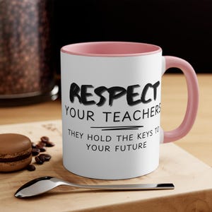 Op de afbeelding: Witte keramische mok met een roze binnenkant en handvat. De mok heeft de tekst "RESPECT YOUR TEACHERS" in het zwart, met de zin "THEY HOLD THE KEYS TO YOUR FUTURE" eronder. Een lepel en een gebakje staan op de voorgrond.