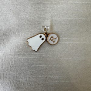 Puede incluir: Un dije de fantasma blanco con ojos negros y contorno dorado, junto a un dije circular con borde dorado con la letra "R" sobre fondo blanco. Ambos dijes están sujetos a un clip de plástico transparente.