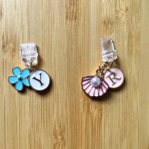 Op de afbeelding: Twee goudkleurige bedels met doorzichtige plastic clips. De eerste bedel heeft een blauwe bloem en een witte cirkel met de letter "Y" in zwart. De tweede bedel heeft een roze schelp met een parel en een roze cirkel met de letter "R" in wit.