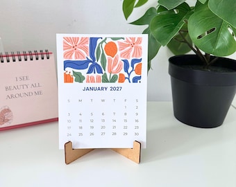 May include: A desk calendar for January 2027 featuring a colorful floral and fruit illustration at the top. The calendar is printed on white paper and is held upright by a small, light brown wooden stand. The design includes pink, blue, and orange.