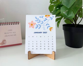 May include: A white desk calendar page for January 2027 featuring a floral illustration with light blue leaves and pink and yellow flowers. The calendar is held upright by a small wooden stand and displays the days of the month in black text.