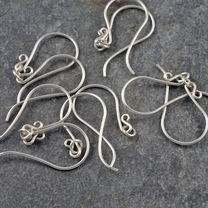 May include: A close-up of six silver earring hooks, all in the shape of a teardrop with a small loop at the top. The hooks are arranged in a random pattern on a grey background.