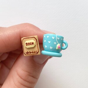 May include: Miniature polymer clay book with the word "BOOK" and a light brown cover. Next to it is a light blue teacup with white heart details and a tea bag. The teacup has a small handle and sits on a matching saucer.