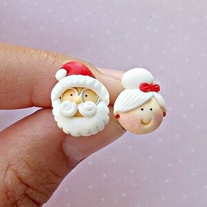 May include: A pair of Christmas-themed earrings featuring Santa Claus and Mrs. Claus. The earrings are made of white polymer clay with red accents. Santa Claus has a white beard and a red hat. Mrs. Claus has a white bun and a red bow.