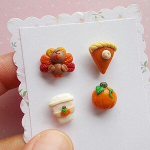May include: A set of four polymer clay earrings featuring Thanksgiving-themed designs: a turkey, a pumpkin, a slice of pumpkin pie, and a coffee cup with a pumpkin on it.