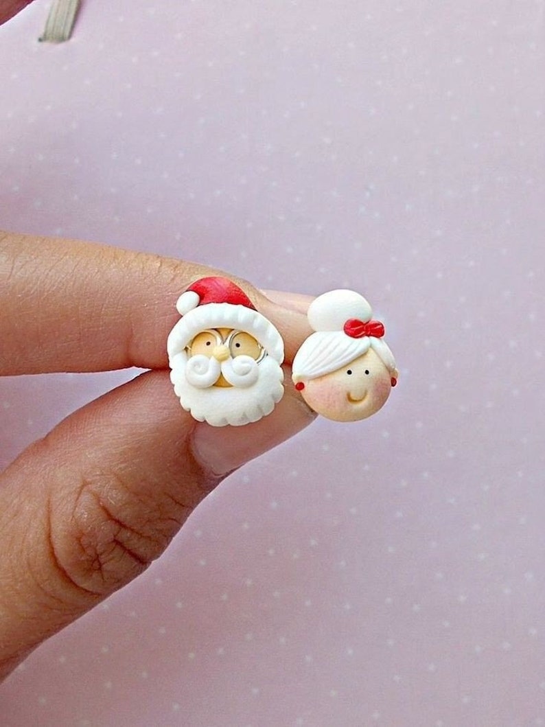 May include: A pair of Christmas-themed earrings featuring Santa Claus and Mrs. Claus. The earrings are made of white polymer clay with red accents. Santa Claus has a white beard and a red hat. Mrs. Claus has a white bun and a red bow.