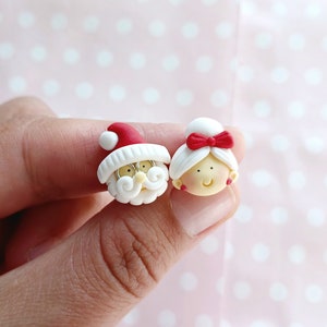 May include: A pair of Christmas-themed earrings featuring a Santa Claus and a Mrs. Claus. The earrings are made of white polymer clay with red accents. Santa Claus has a white beard and a red hat. Mrs. Claus has a red bow in her hair.