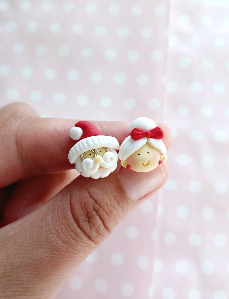 May include: A pair of Christmas-themed earrings featuring a Santa Claus and a Mrs. Claus. The earrings are made of white polymer clay with red accents. Santa Claus has a white beard and a red hat. Mrs. Claus has a red bow in her hair.