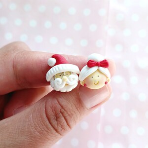 May include: A pair of white clay earrings with red accents. One earring features a Santa Claus face with a white beard and a red hat. The other earring features a woman's face with a red bow in her hair.