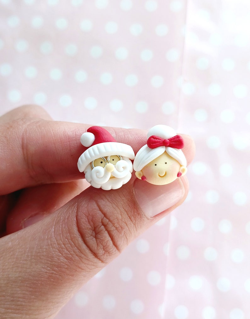 May include: A pair of white clay earrings with red accents. One earring features a Santa Claus face with a white beard and a red hat. The other earring features a woman's face with a red bow in her hair.