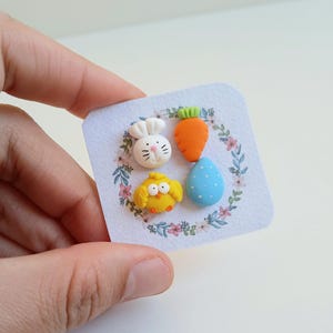 May include: A set of four Easter-themed polymer clay earrings. The earrings feature a white bunny, a yellow chick, an orange carrot, and a blue egg with white polka dots.