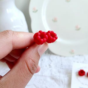 以下が含まれることがあります： 明るい赤色のラズベリー型のスタッドピアス。ピアスは手で持たれており、背景には花のアクセントが付いた白いプレートがあります。ピアスは小さく、詳細で、質感のある表面です。